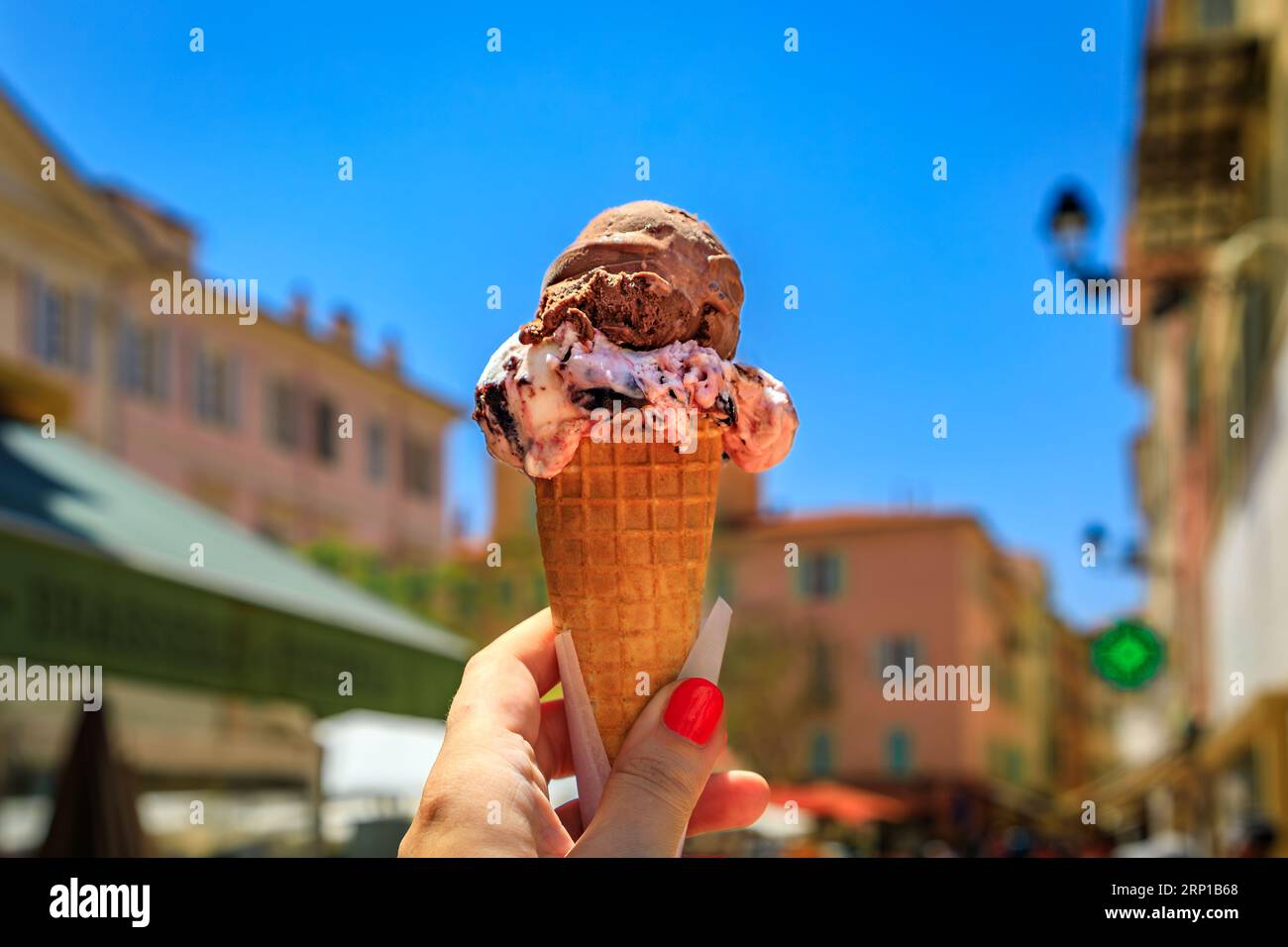 Main de femme tenant un gelato au chocolat artisanal dans un cône, vue sur les maisons colorées traditionnelles dans la vieille ville Vieille ville de Menton Sud de la France Banque D'Images