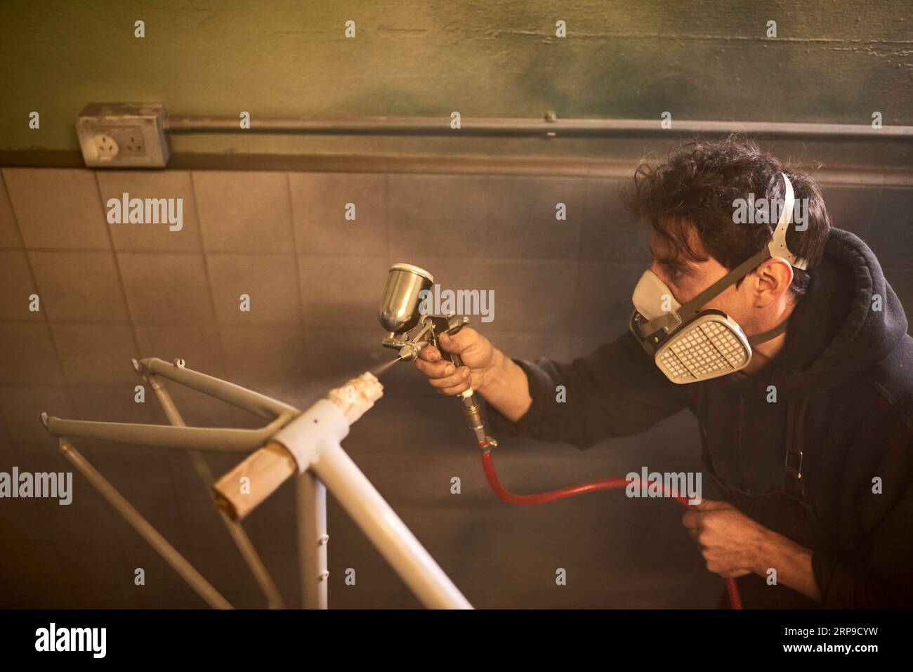 Jeune homme hispanique portant un masque de protection concentré peinture au pistolet un cadre de bicyclette dans son atelier. Composition avec espace de copie. Banque D'Images