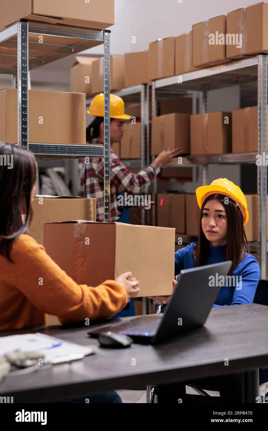Responsable d'entrepôt supervisant l'expédition des produits et l'envoi des colis. L'opérateur de magasin prend la boîte en carton d'un collègue asiatique et coordonne la qualité de l'emballage des commandes Banque D'Images