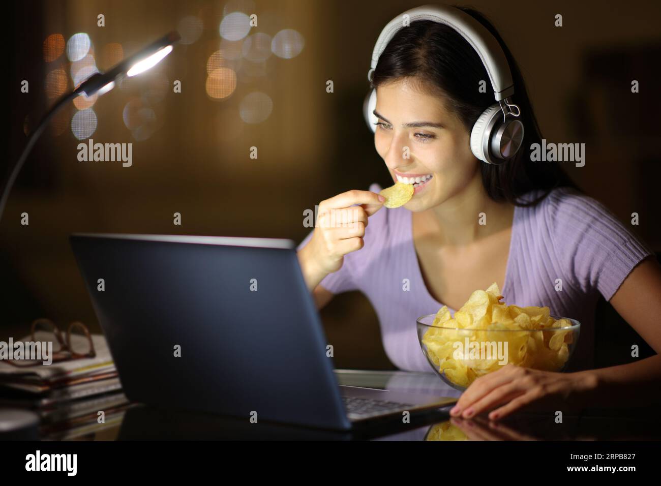 Femme heureuse dans la nuit portant un casque mangeant des chips de pomme de terre regarder des médias sur ordinateur portable à la maison Banque D'Images