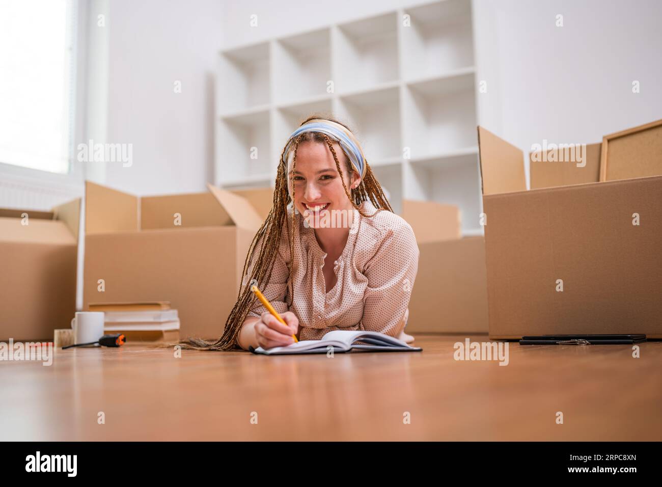 Femme au gingembre moderne avec des tresses écrivant dans un cahier tout en déménageant dans une nouvelle maison. Banque D'Images