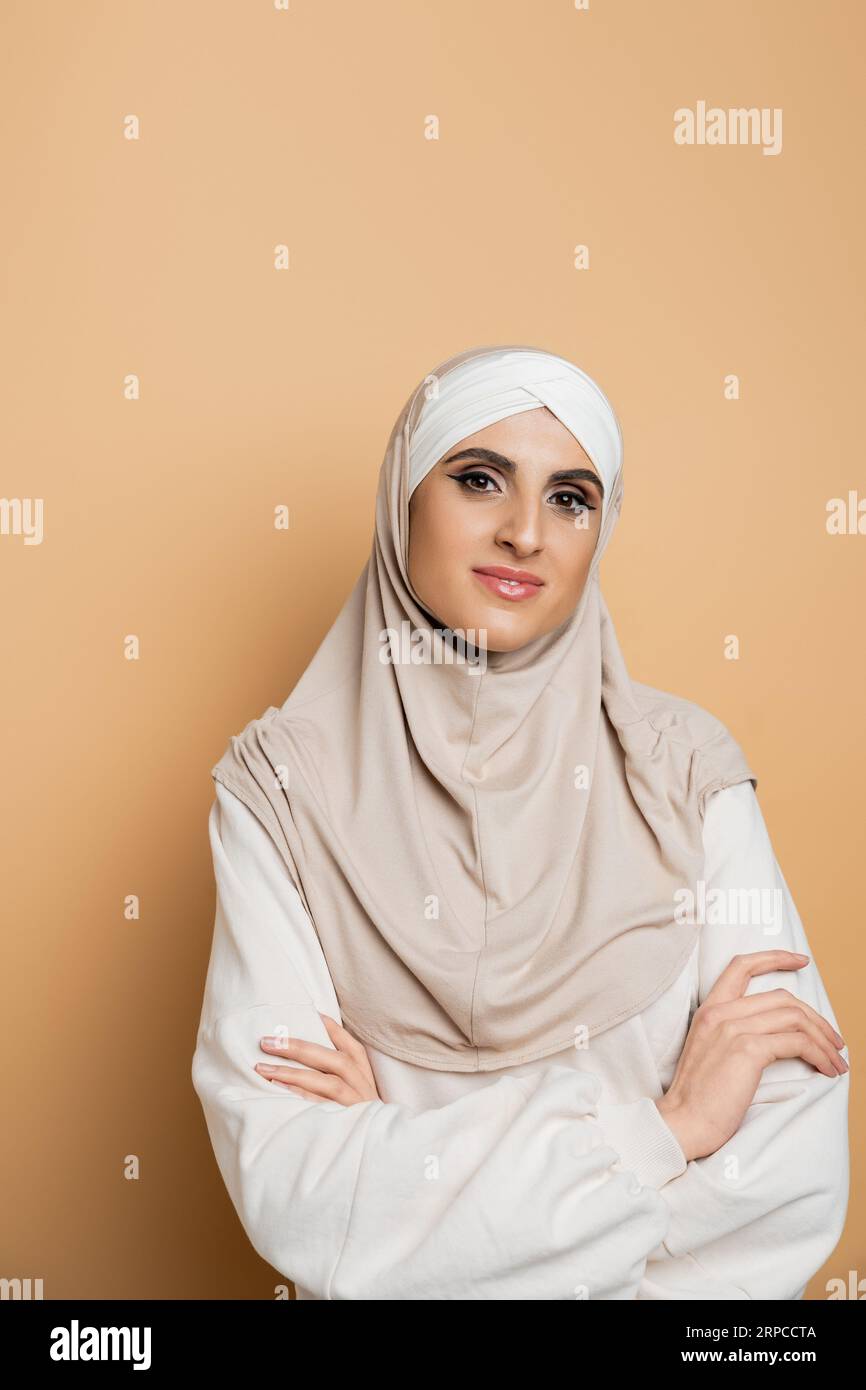 femme musulmane confiante en hijab et sweat-shirt posant avec les bras pliés, souriant à la caméra sur beige Banque D'Images