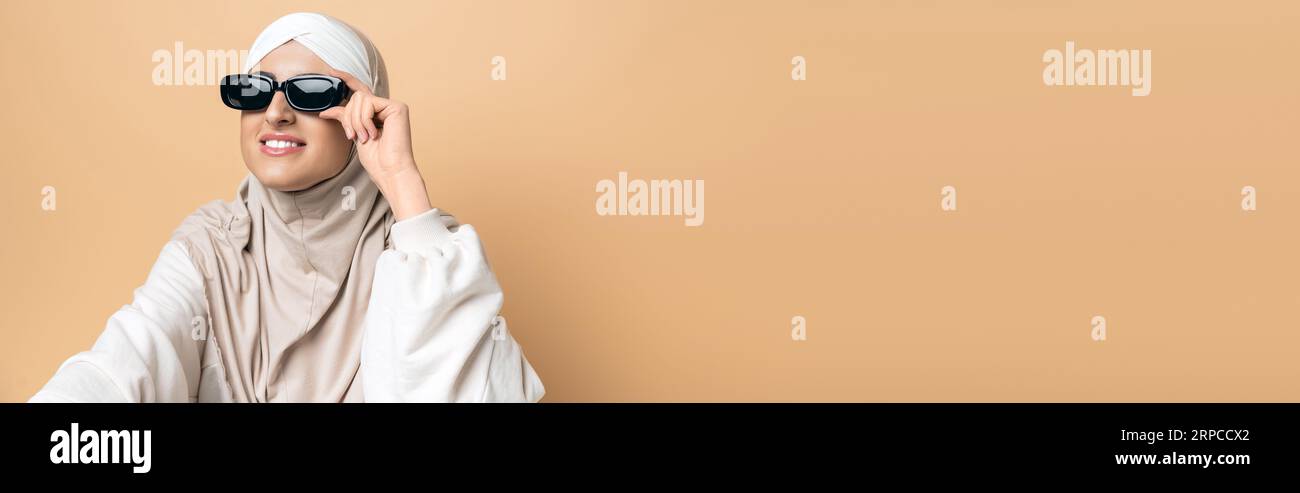 femme musulmane joyeuse en hijab et lunettes de soleil à la mode regardant loin sur beige, style personnel, bannière Banque D'Images