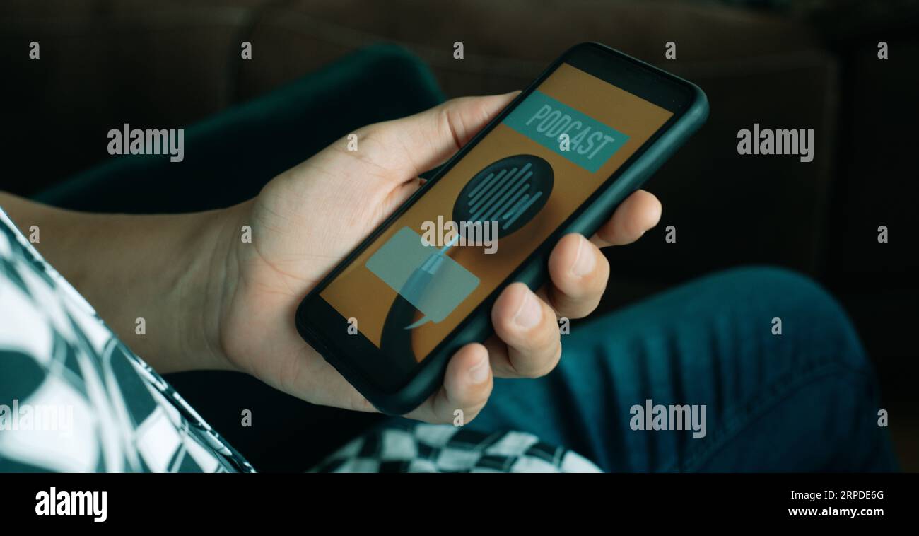 gros plan un jeune homme, en tenue décontractée, assis dans une chaise confortable, avec son smartphone à la main, qui lit le texte podcast sur son écran Banque D'Images