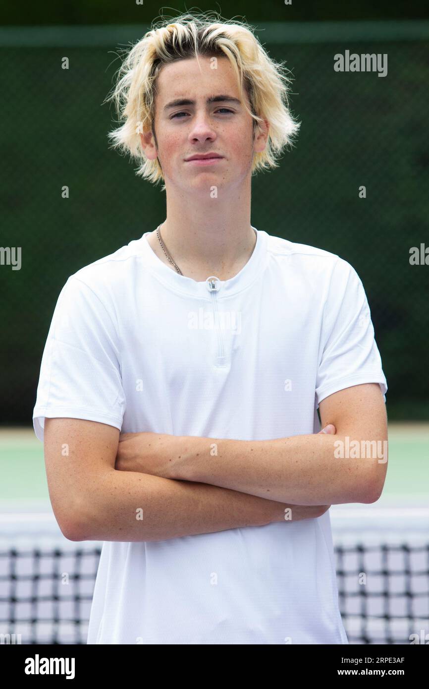 Jeune joueur de tennis caucasien déterminé pose pour la caméra dans un t-shirt blanc et un short rouge, affichant leur posture athlétique concentrée. Banque D'Images