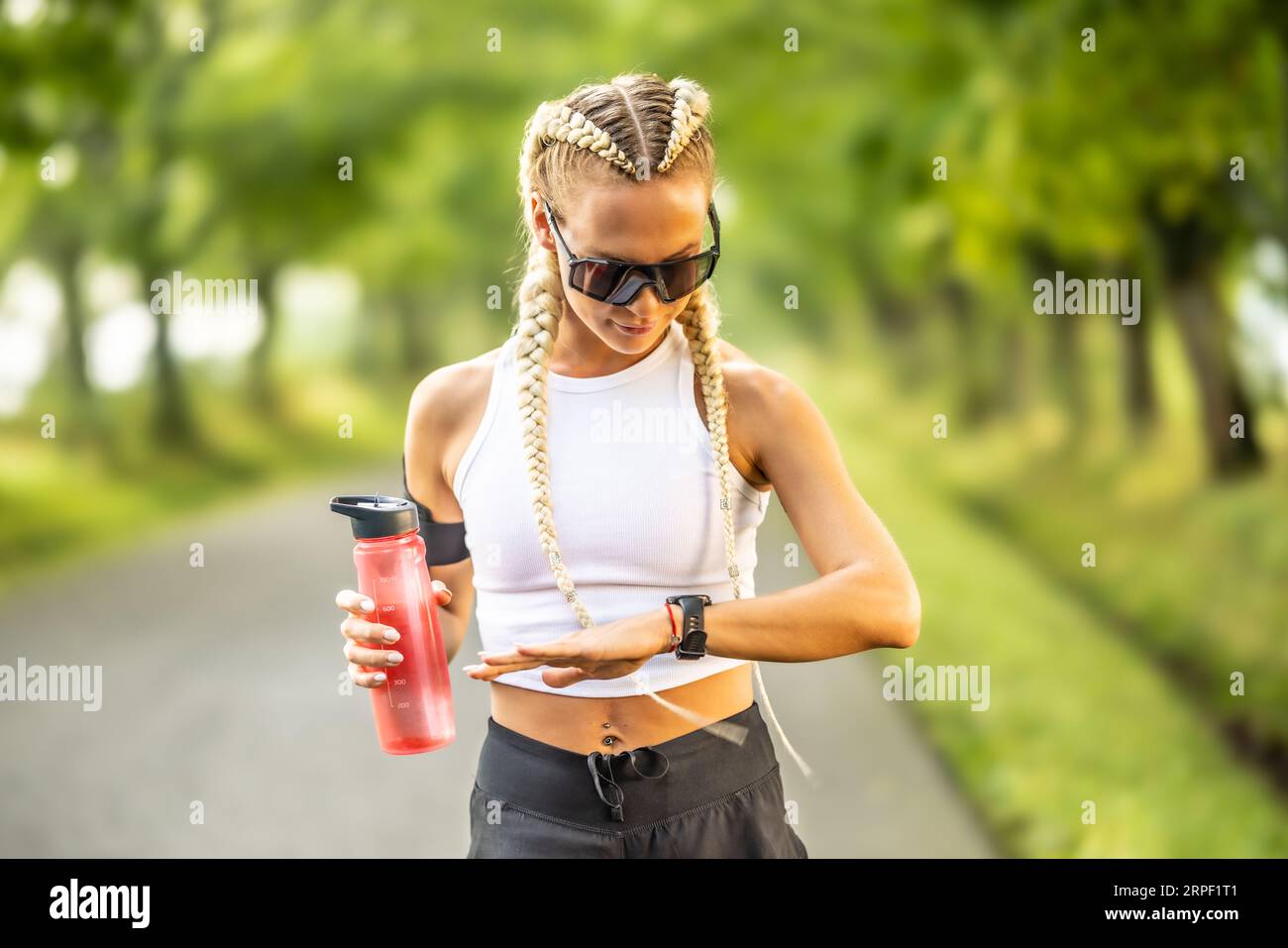 Belle athlète féminine vérifiant sa fréquence cardiaque sur la montre intelligente, tenant la bouteille d'eau pour l'hydratation avant le marathon. Banque D'Images