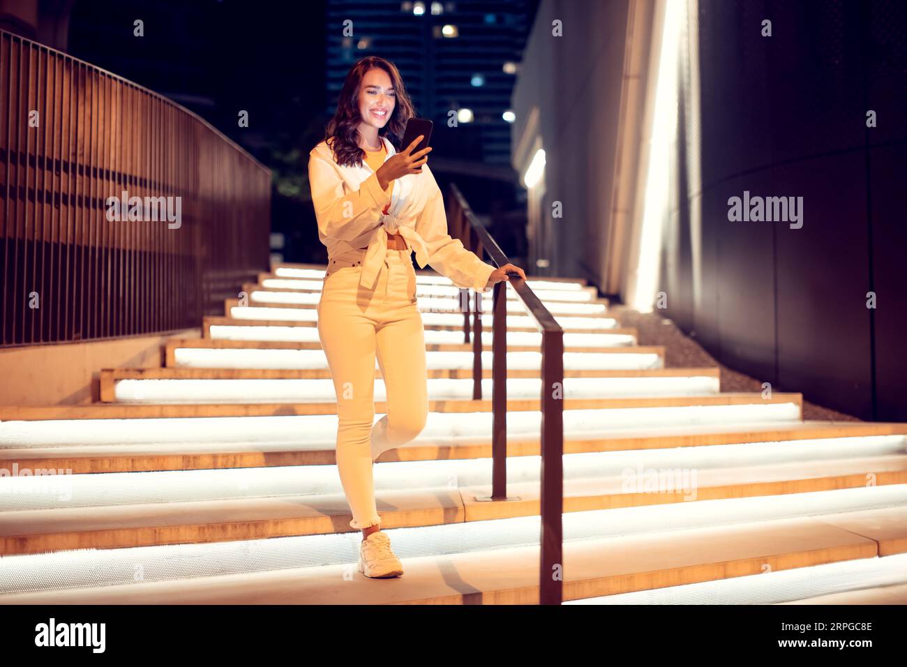 Jeune femme utilisant un téléphone portable pendant la promenade dans la ville de nuit, en descendant les escaliers illuminés, espace de copie, pleine longueur Banque D'Images