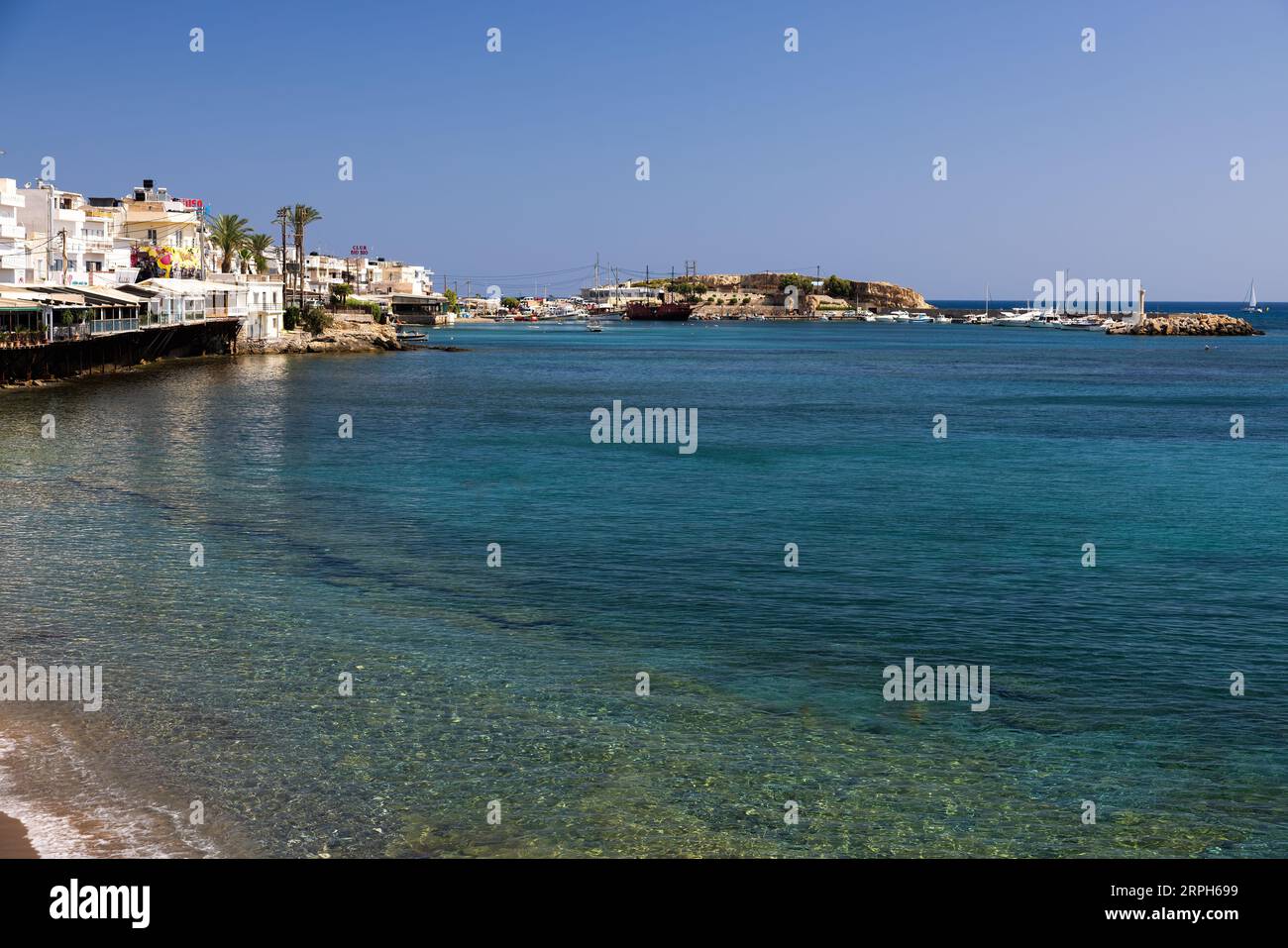 Prélassez-vous dans l'étreinte du soleil tout en admirant le rivage animé et le port animé d'Hersonissos. Sur la gauche, des restaurants pittoresques ornent la scène, tandis que les navires Banque D'Images