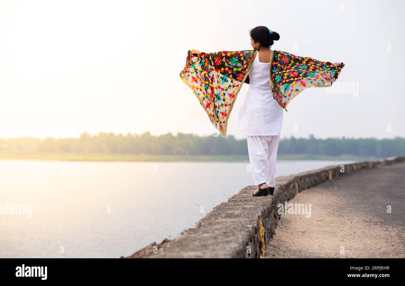 Une jeune fille indienne en robe blanche et colorée marchant le long d'une rivière et profitant du vent avec les mains tendues Banque D'Images