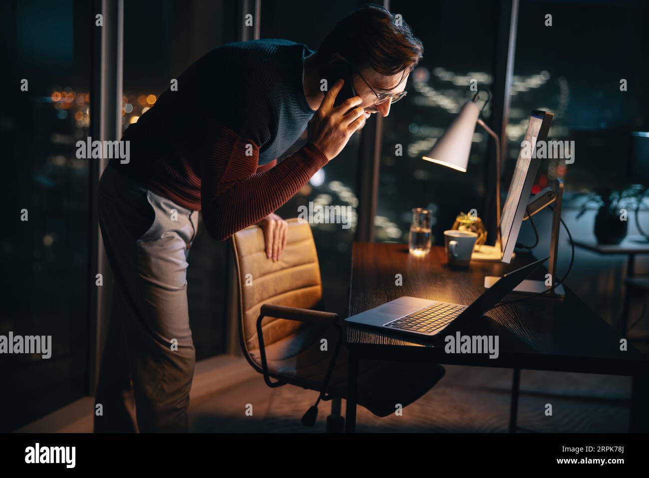 Un homme d'affaires travaillant tard le soir dans un bureau. Il discute d'un projet au cours d'un appel téléphonique tout en utilisant un ordinateur portable et un téléphone portable. Dédié Banque D'Images