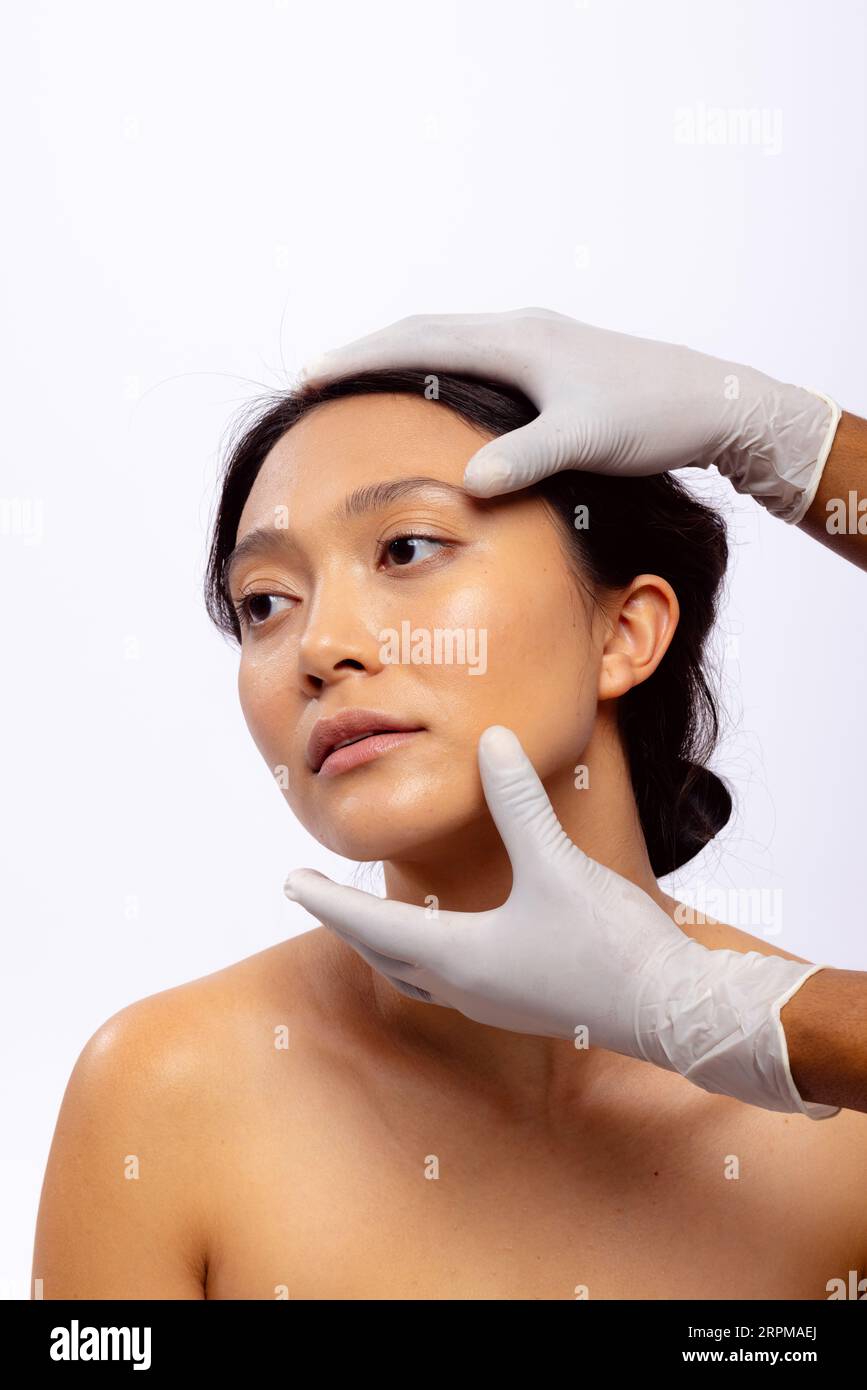 Mains de l'esthéticienne dans des gants chirurgicaux blancs tenant le visage de la femme asiatique avec les cheveux foncés, espace de copie Banque D'Images