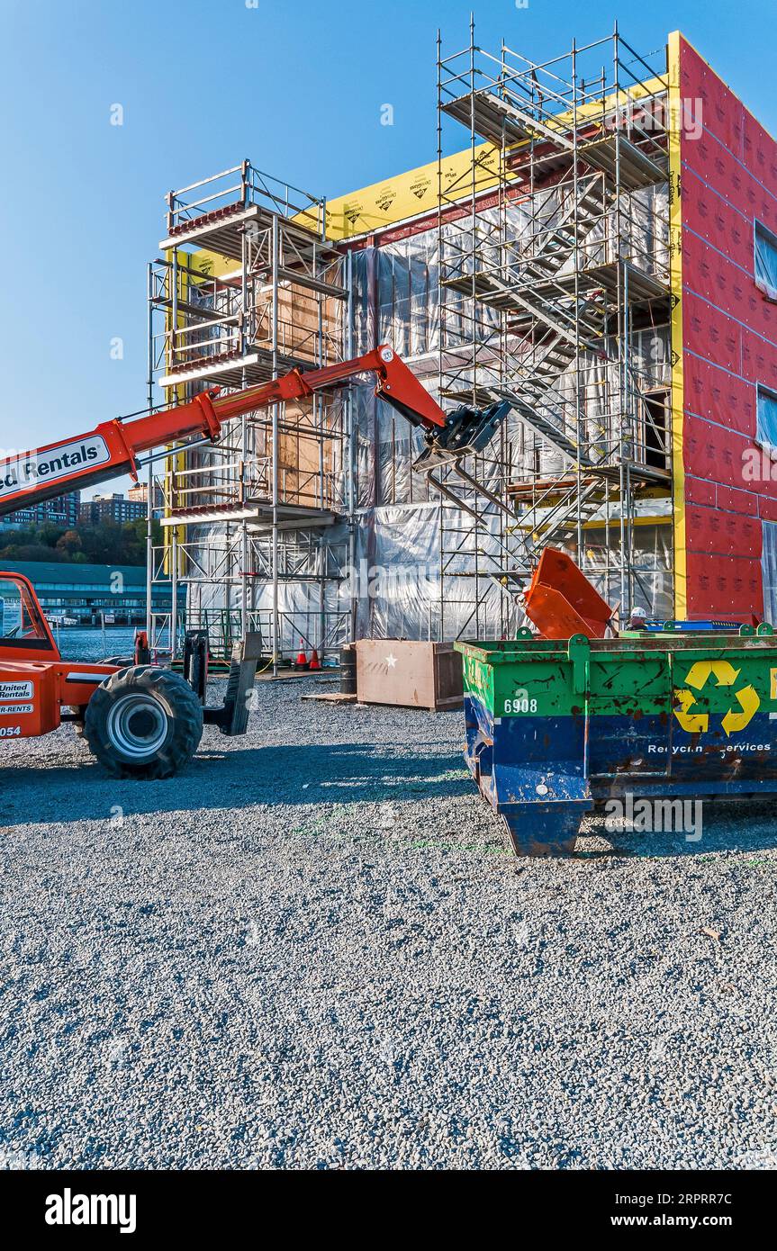 Un haut-ascenseur de location orange avec son bras étendu au toit d'un bâtiment sur un chantier de construction, tandis qu'un travailleur de la construction dans un casque de dur marche sur le site. Banque D'Images