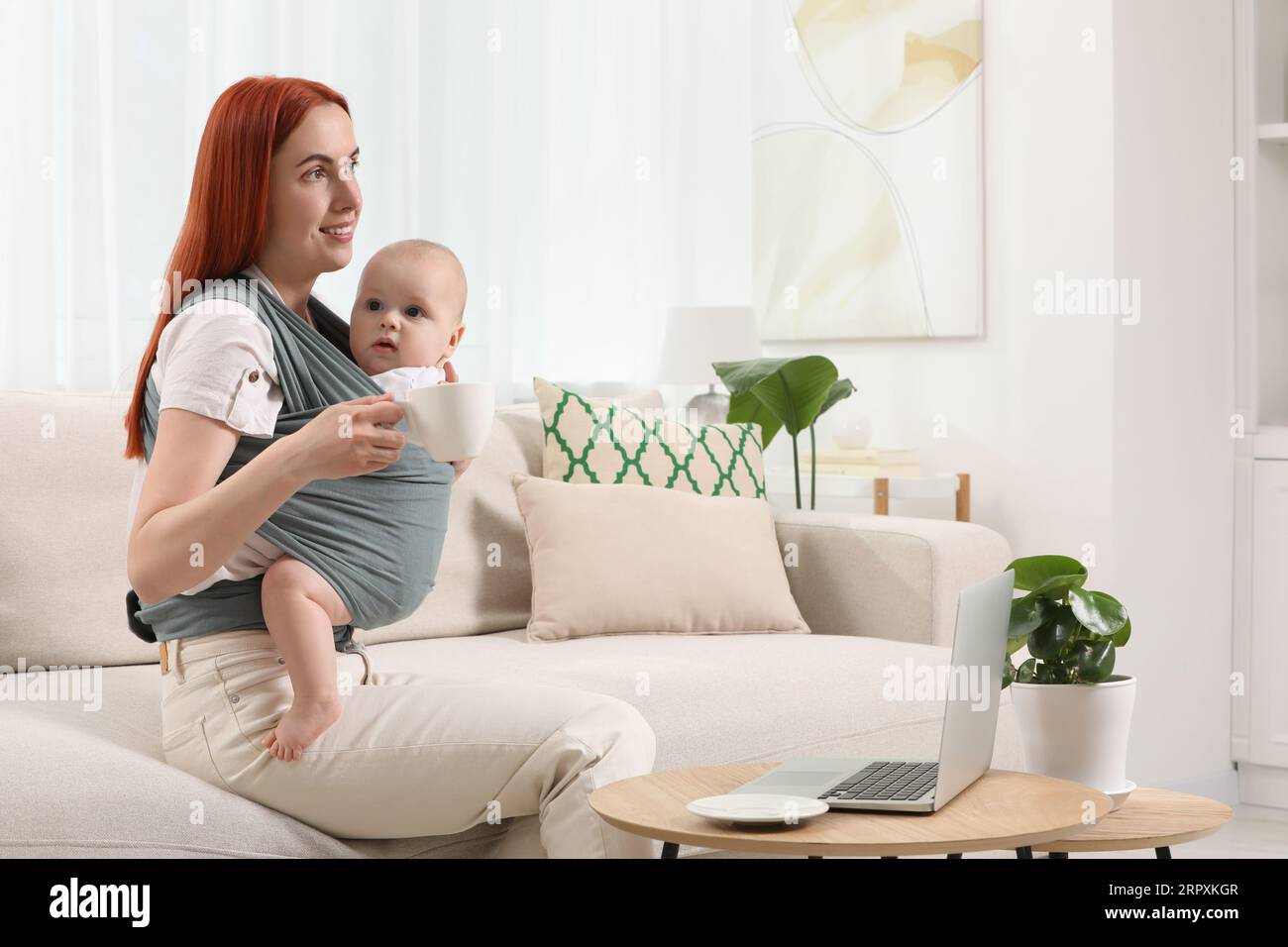 Mère avec une tasse de boisson tenant son enfant en fronde (porte-bébé) à la maison Banque D'Images