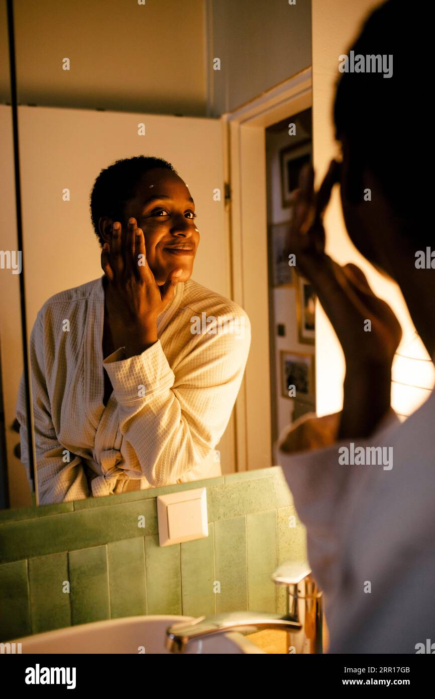 Jeune femme souriante appliquant une crème hydratante sur le visage tout en regardant dans le miroir à la maison Banque D'Images