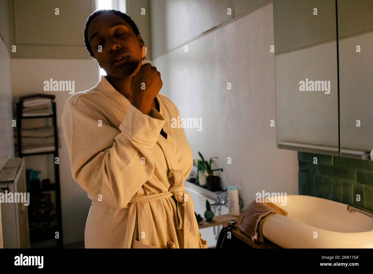 Femme avec les yeux fermés en utilisant le rouleau de jade sur le visage dans la salle de bain à la maison Banque D'Images