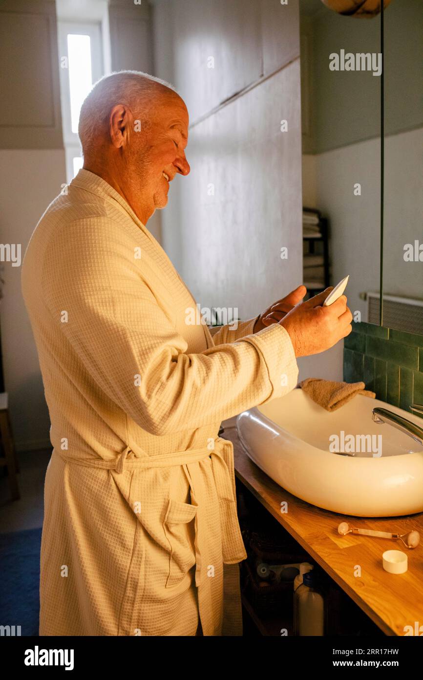 Homme âgé souriant tout en appliquant un hydratant dans la salle de bain à la maison Banque D'Images
