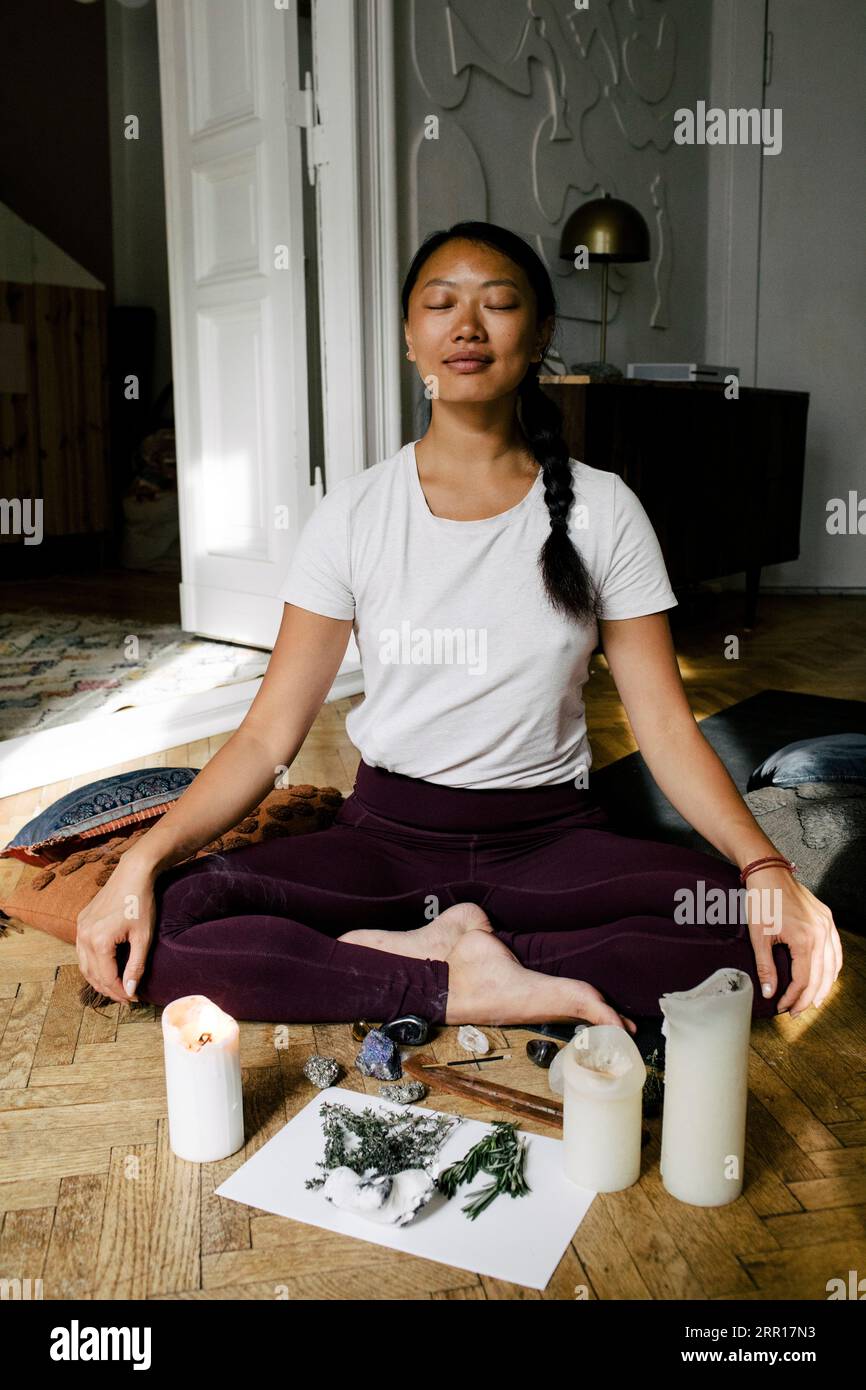 Jeune femme souriante méditant devant des herbes et allumant des bougies à la maison Banque D'Images