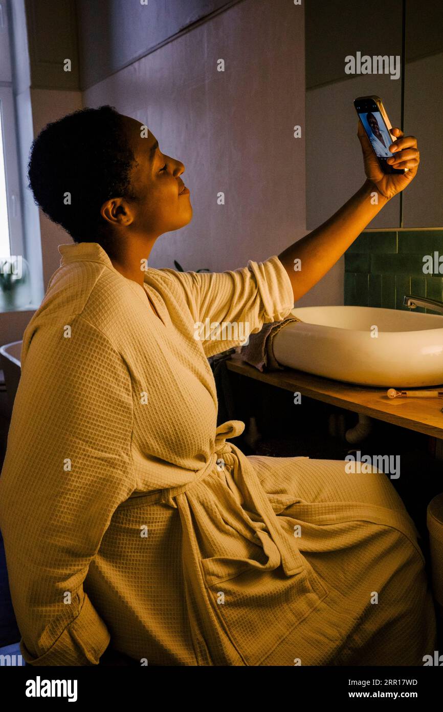 Femme souriante prenant selfie par téléphone intelligent tout en étant assis dans la salle de bain Banque D'Images