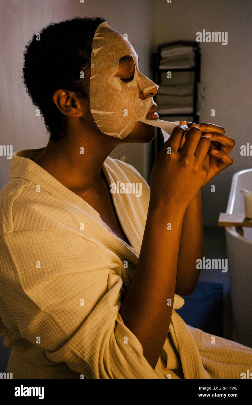 Jeune femme enlevant le masque facial sur le visage tout en étant assise dans la salle de bain Banque D'Images