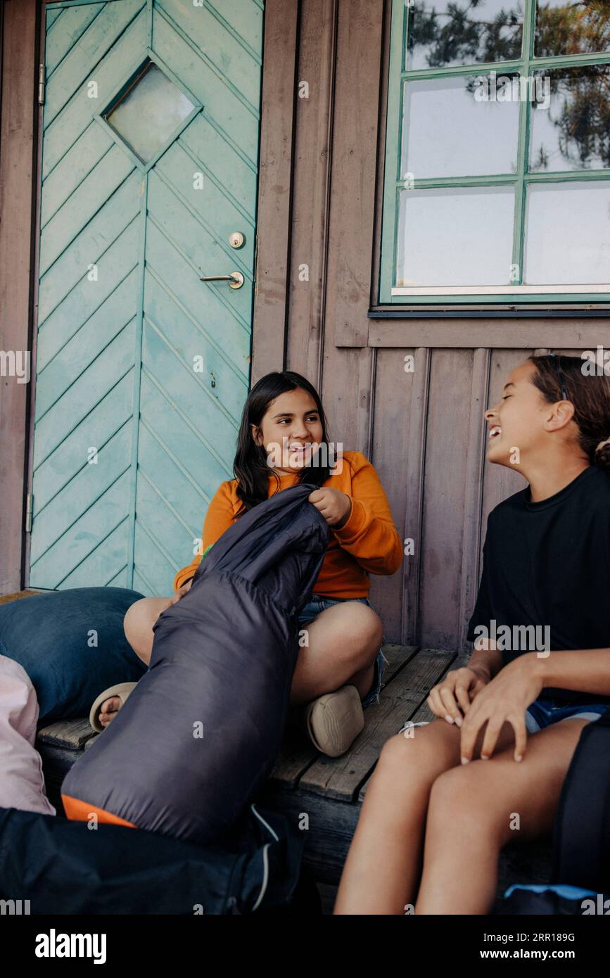 Heureux amis féminins assis avec des sacs à dos sur le porche à l'extérieur de la cabine au camp d'été Banque D'Images