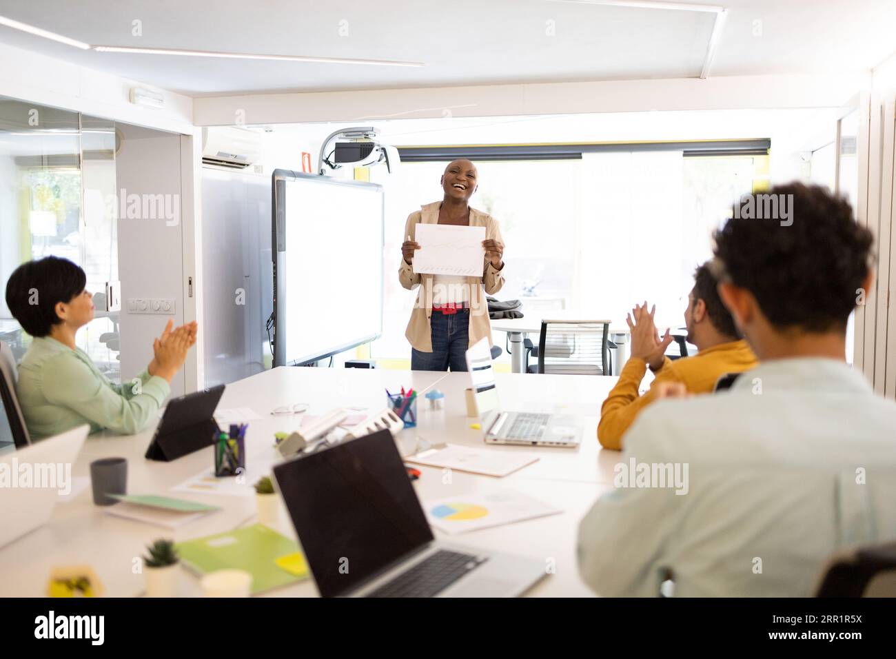 Femme afro-américaine montrant la feuille de papier blanc à des collègues applaudissant les mains et assis au bureau blanc dans le bureau moderne Banque D'Images