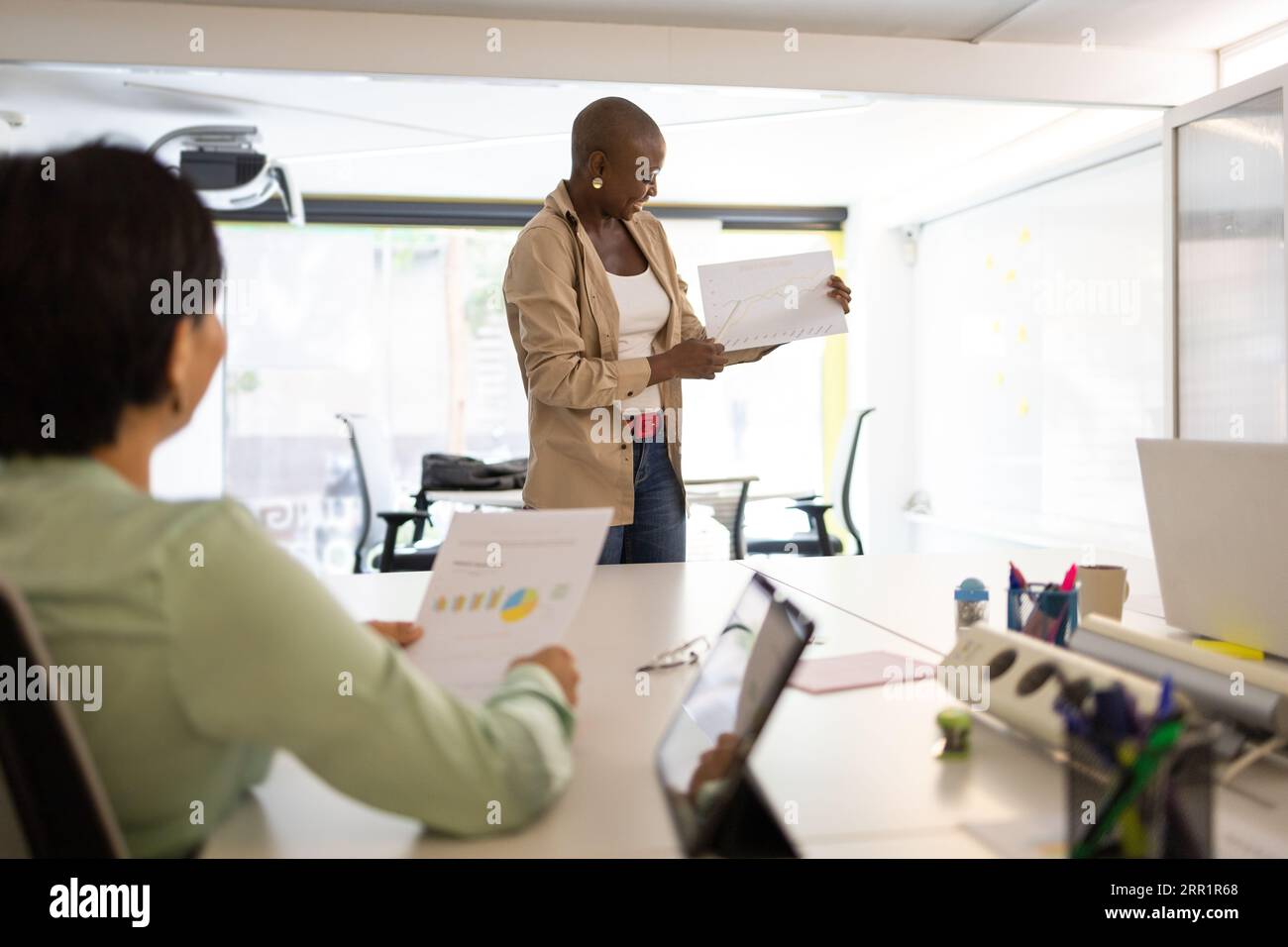 Femme afro-américaine montrant une feuille de papier blanc à un collègue assis à un bureau blanc dans un bureau moderne Banque D'Images