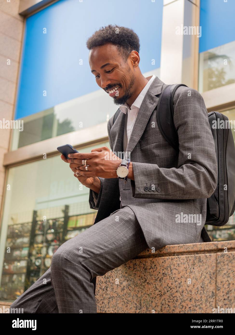 Homme afro-américain ravi dans des vêtements formels assis sur le banc de rue en pierre et naviguant téléphone portable dans la lumière du soleil Banque D'Images