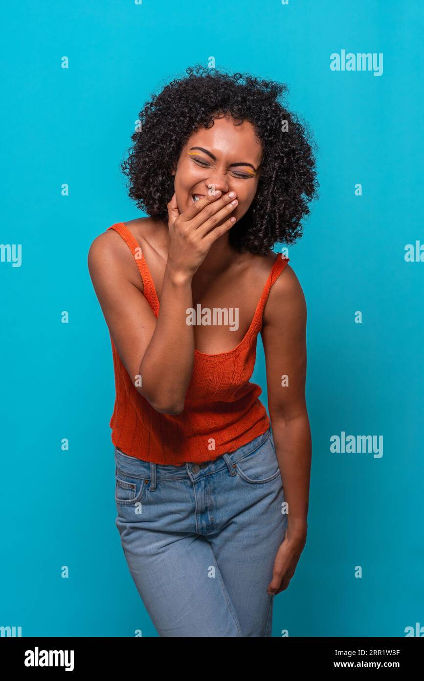 Modèle féminin afro-américain excité avec coiffure afro en tenue décontractée riant à la blague avec les yeux fermés et couvrant le visage sur fond bleu Banque D'Images