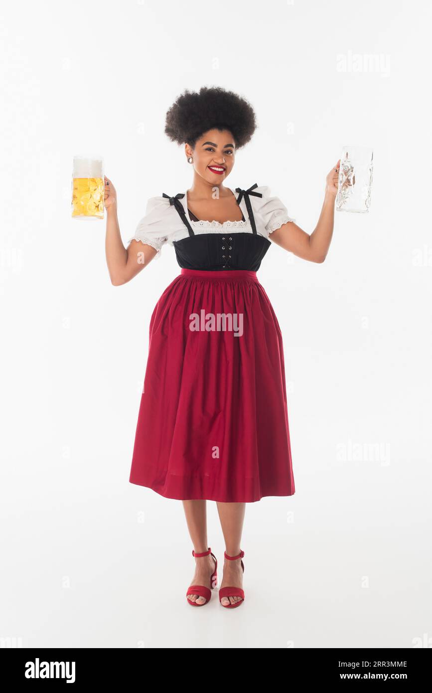 serveuse afro-américaine oktoberfest en costume traditionnel avec des tasses à bière vides et pleines sur blanc Banque D'Images