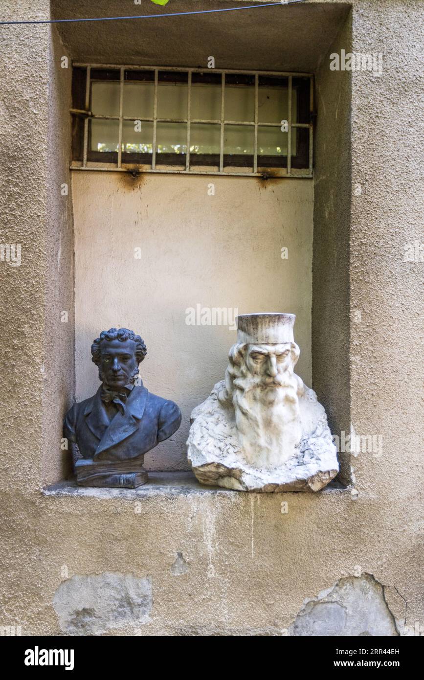 Deux bustes se dressent dans une niche murale sous une fenêtre grillagée sur un bâtiment de la rue Gia Chanturia dans le quartier Sololaki de Tbilissi, en Géorgie. La buste sombre à gauche représente un jeune homme aux cheveux bouclés, aux favoris, portant un nœud papillon et un manteau (peut-être le poète Alexandre Pouchkine). La buste blanche à droite représente un homme à la longue barbe et au couvre-chef cylindrique. Le plâtre rugueux du mur s’est écaillé par endroits. Banque D'Images