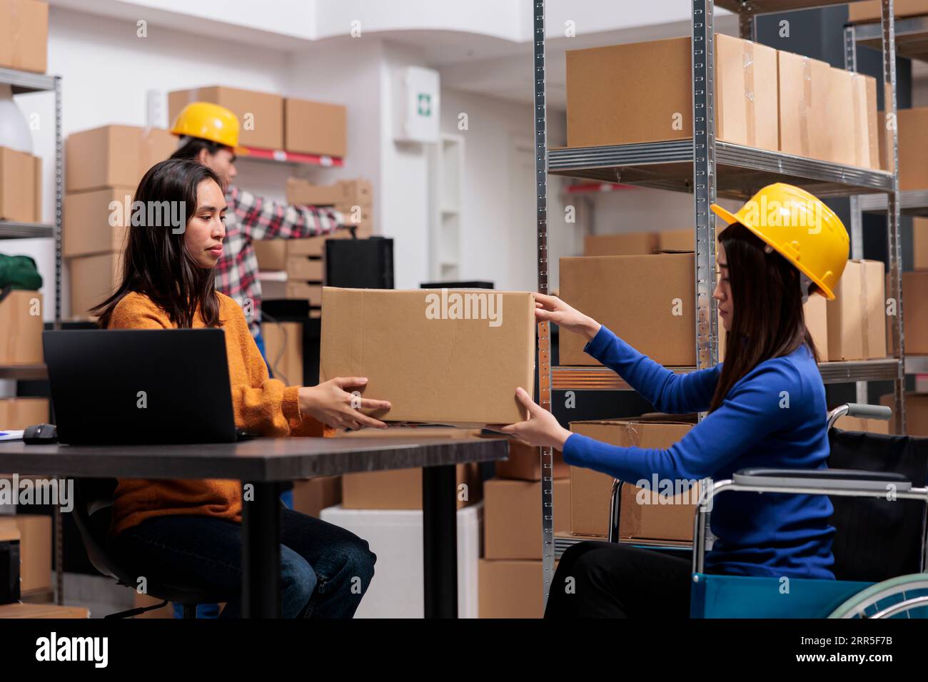 Préparateur de commande en fauteuil roulant donnant le colis à l'opérateur de livraison dans l'entrepôt. Responsable de la distribution assis à son bureau et prenant la boîte en carton de l'employé de l'entrepôt pour préparer le produit à expédier Banque D'Images