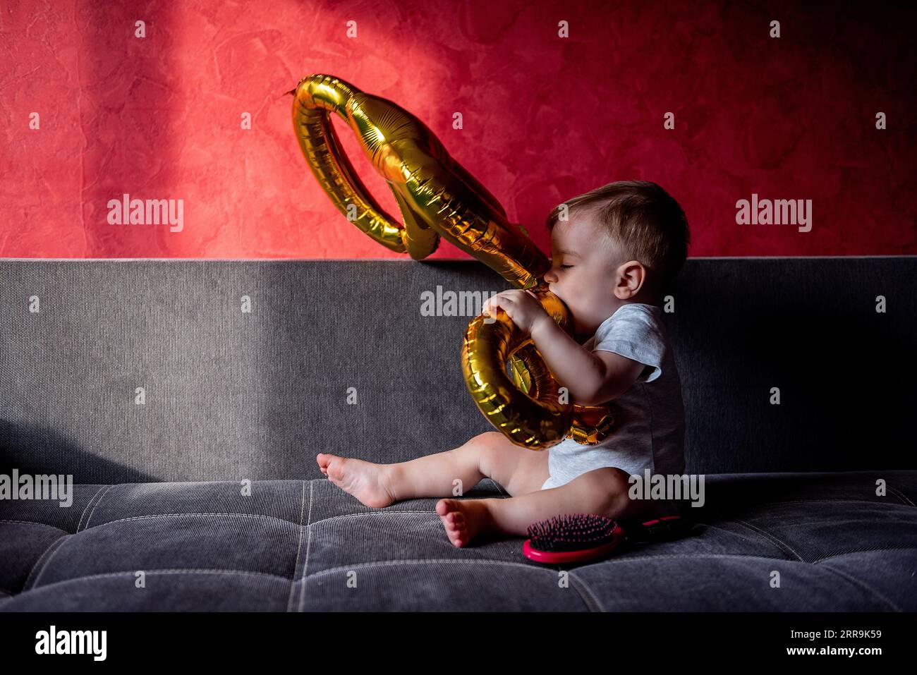 Première fête d'anniversaire, bébé joue avec des ballons d'aluminium de couleur or UN. Petit garçon se trompant, assis sur un canapé gris au soleil. Dessin ombre claire activé Banque D'Images