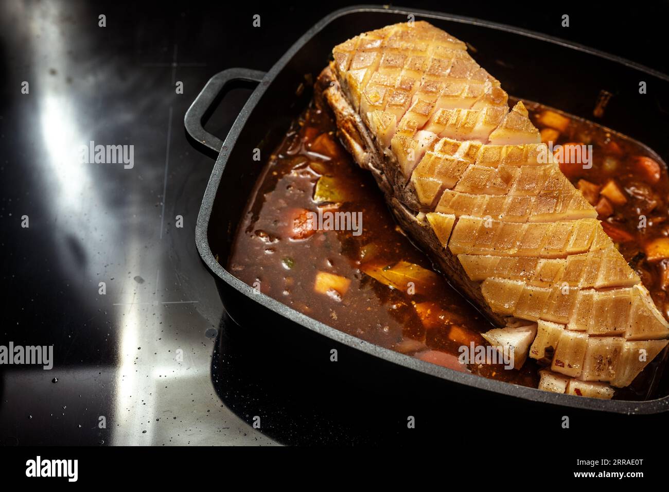Morceau de viande de porc avec croûte grasse incisée sur une base de sauce brune de légumes et de vin rouge dans une poêle noire sur le poêle préparé pour un delicio Banque D'Images