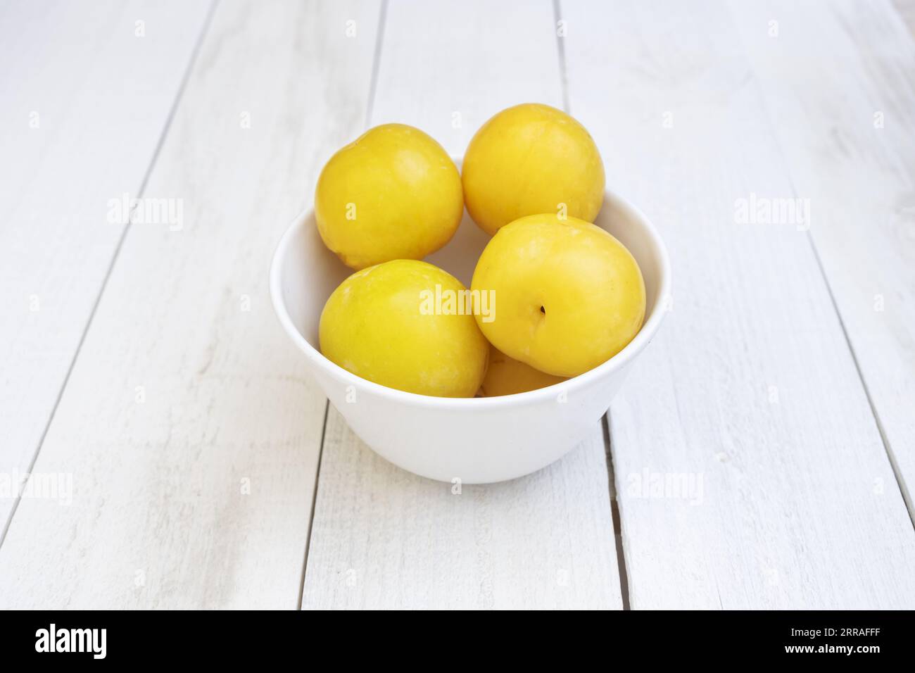 Un petit bol en porcelaine blanche rempli de prunes jaunes mûres sur une table en bois blanc Banque D'Images