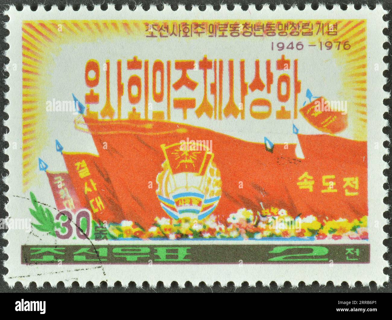 Timbre-poste annulé imprimé par la Corée du Nord, qui montre les armoiries et le drapeau, le 30e anniversaire de la jeunesse ouvrière socialiste, vers 1976. Banque D'Images