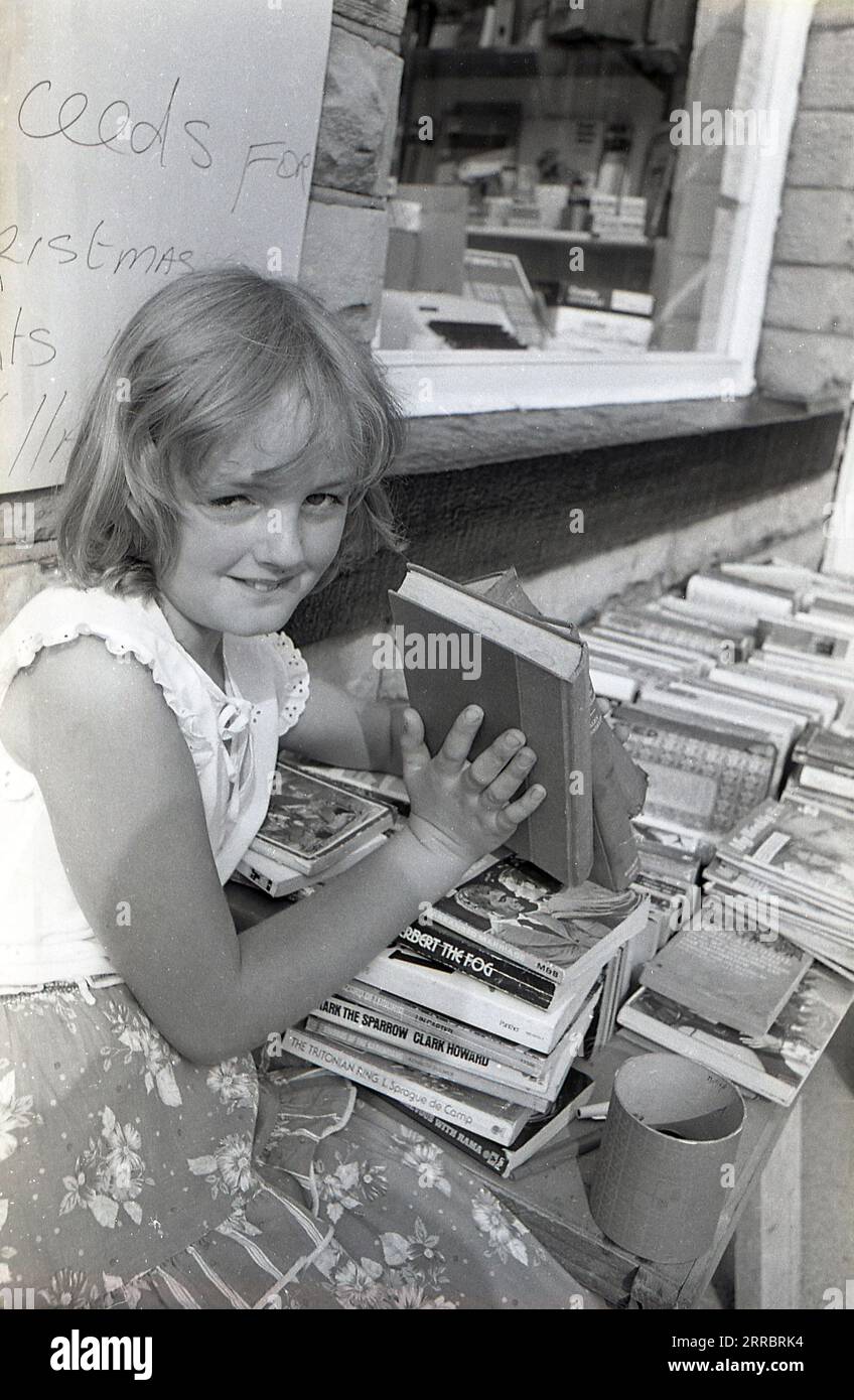 1983, historique, une jeune fille vendant des livres d'occasion à partir d'un étal devant un magasin de papeterie viilage, Yorkshire, Angleterre, Royaume-Uni pour essayer de collecter des fonds pour des lumières de noël dans son village. Banque D'Images