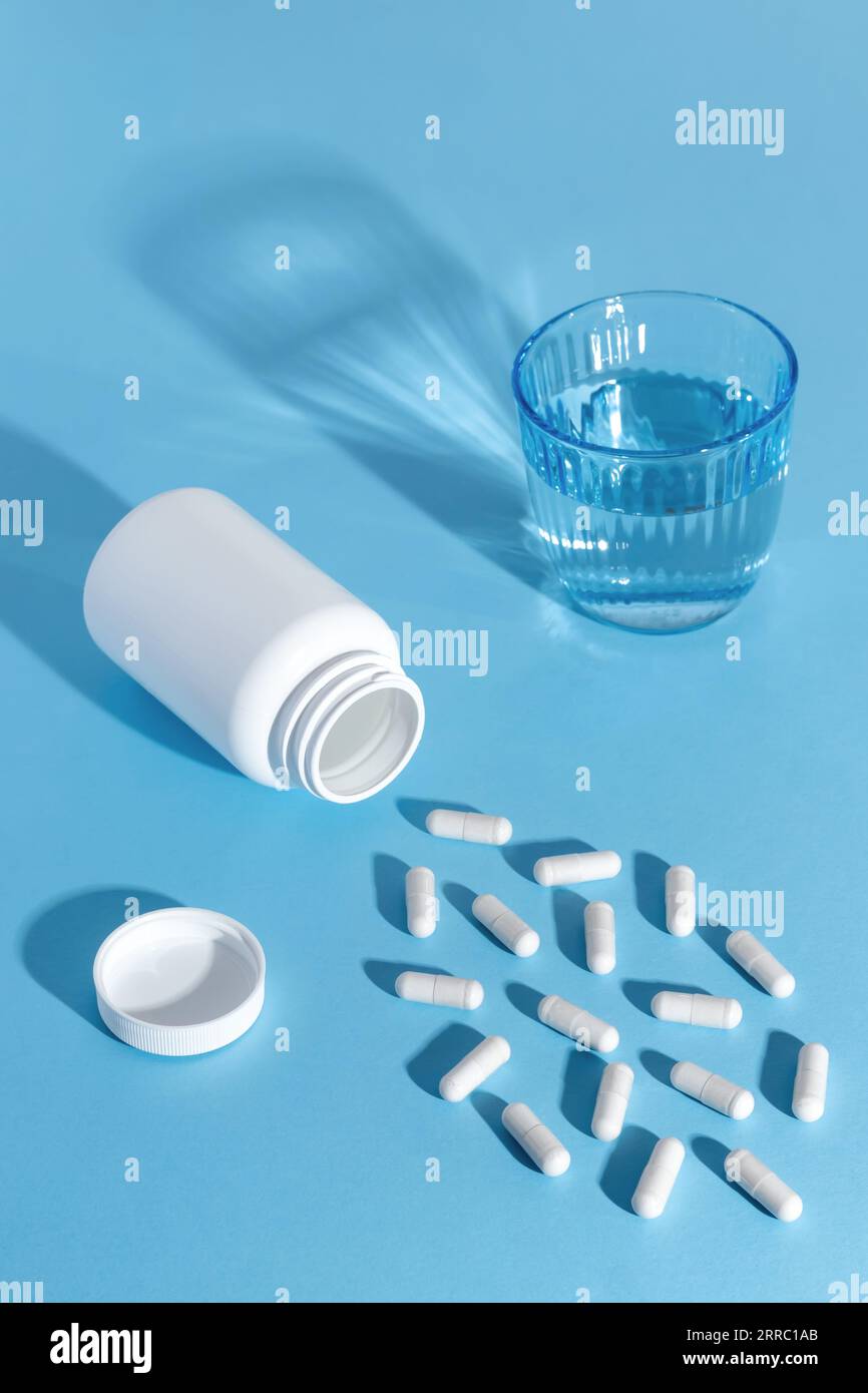 Pilules médicales blanches, capsules et verre d'eau sur fond bleu. Nature morte avec des ombres dures. Un tas de comprimés éparpillés. Concept de soins de santé. Banque D'Images
