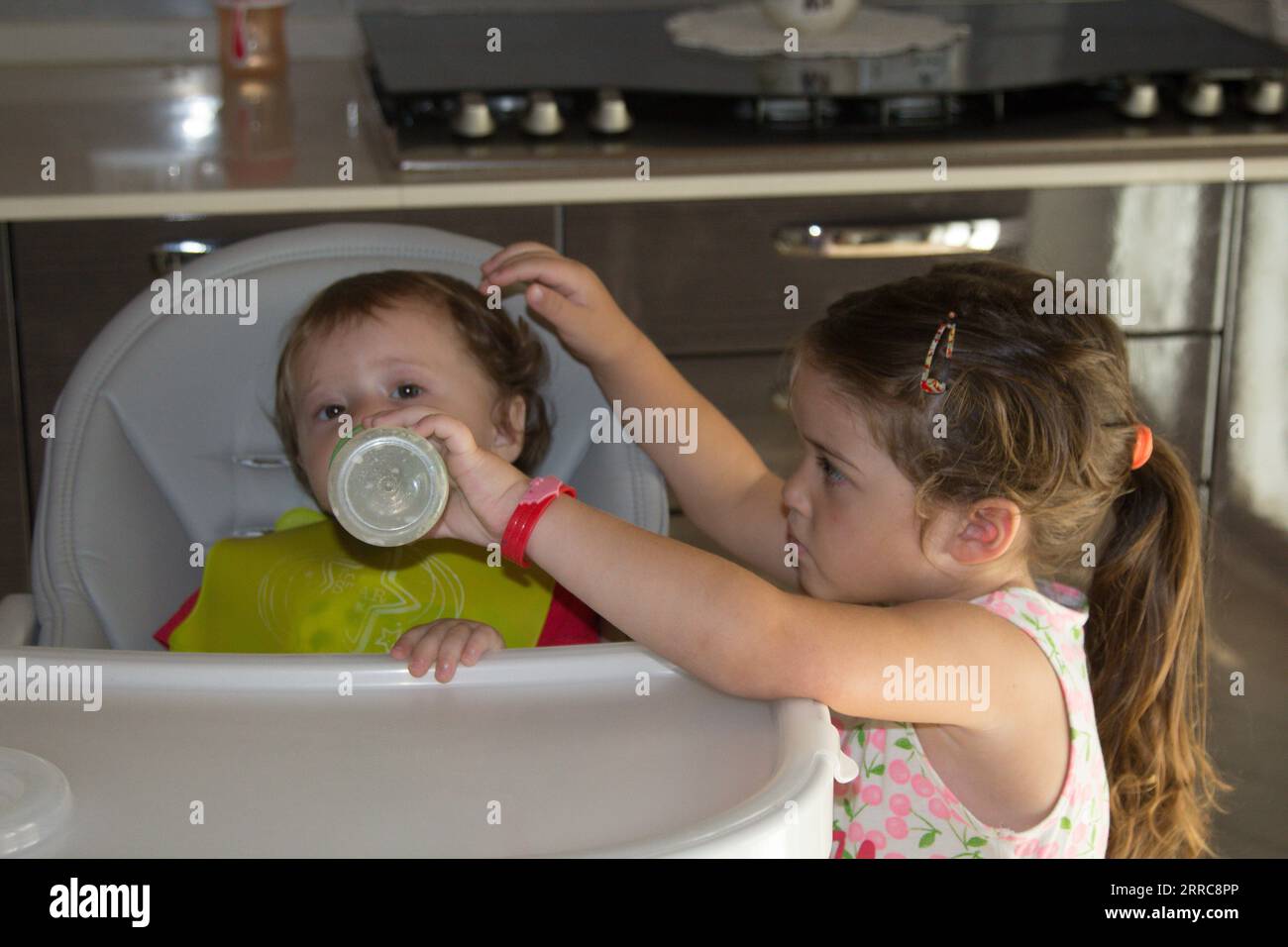 Image d'une adorable petite fille donnant du lait à son jeune frère dans la chaise haute avec une bouteille. Amour fraternel Banque D'Images