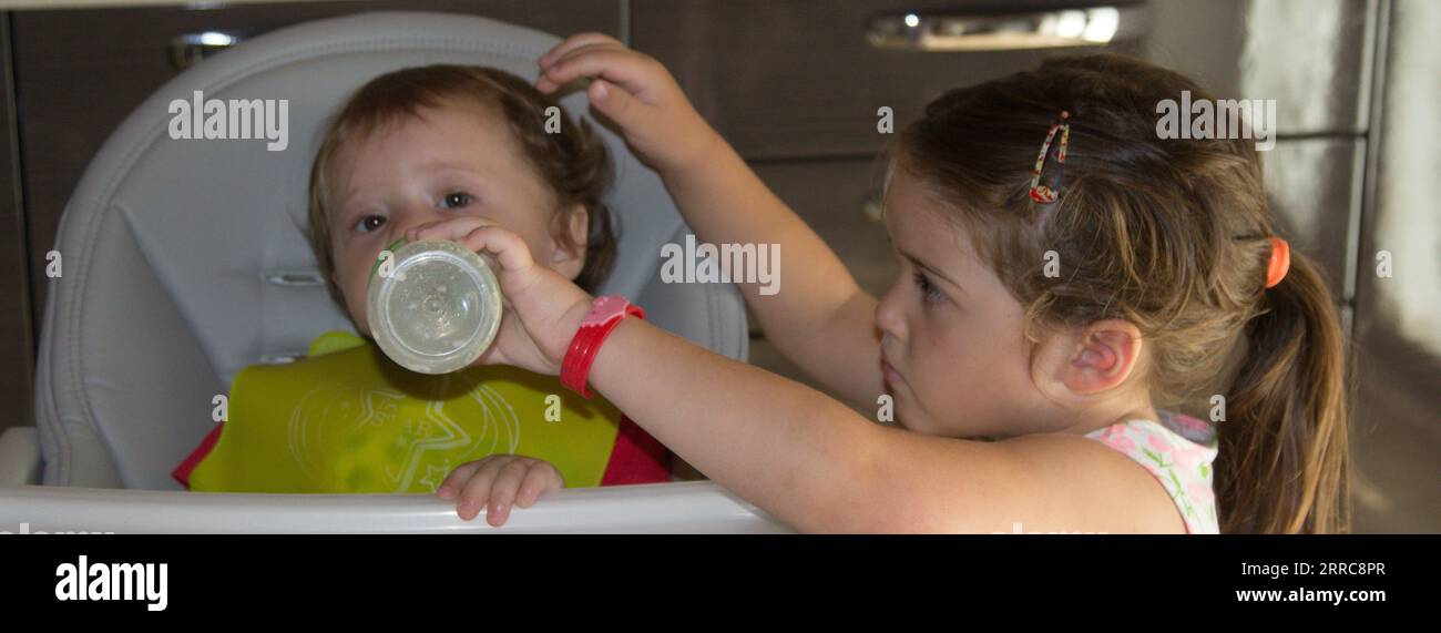 Image d'une adorable petite fille donnant du lait à son jeune frère dans la chaise haute avec une bouteille. Amour fraternel. Bannière horizontale Banque D'Images