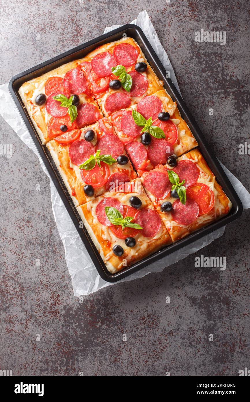 Pizza rectangulaire avec fromage, saucisse au salami, olives et tomates en gros plan dans une plaque de cuisson sur la table. Vue verticale de dessus Banque D'Images