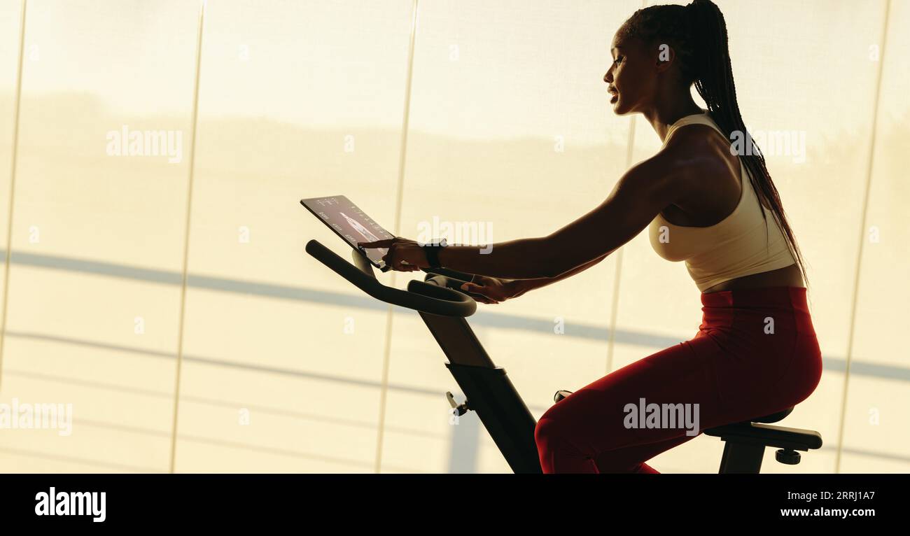 Jeune femme africaine s'exerçant sur un vélo d'exercice intelligent avec une tablette tactile. Elle participe à un cours de fitness numérique à la maison, en train de travailler Banque D'Images