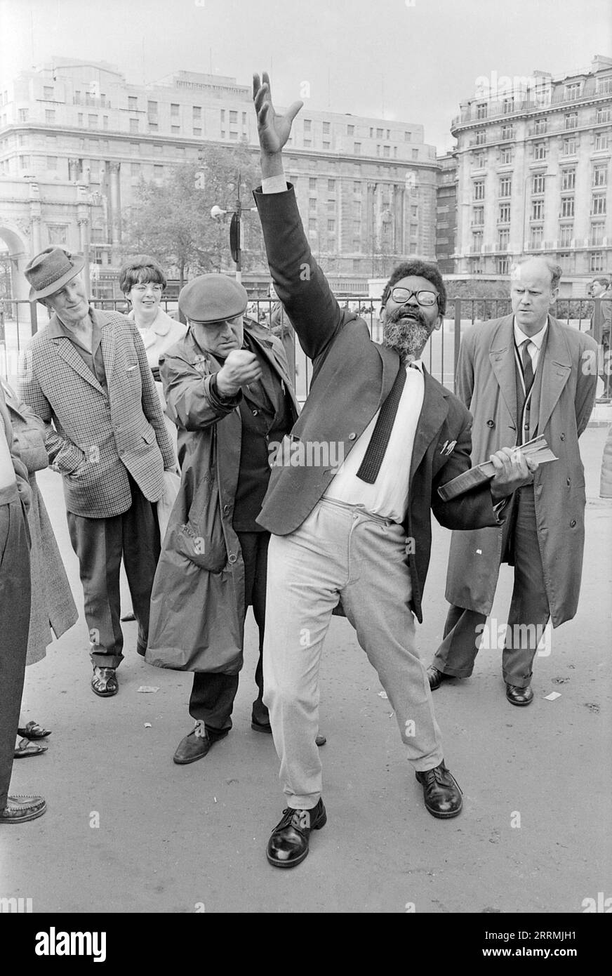 Londres. 1965 – évangéliste prêchant à Speaker’s Corner, Hyde Park, à la jonction de Cumberland Gate, près de Marble Arch, Londres. Le prédicateur tient une bible dans une main et lève son autre main en l'air. Un groupe de spectateurs se sont rassemblés autour de lui. Un homme âgé portant un imperméable et une casquette plate et serrant sa main dans un poing, se moquant du prédicateur. L'hôtel Cumberland et le bâtiment situé au 140 Park Lane sont visibles au loin. Banque D'Images