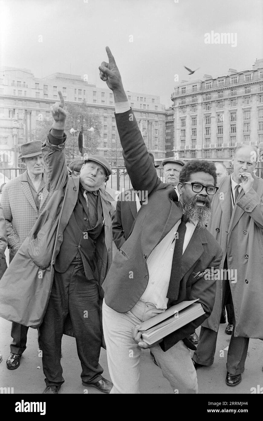 Londres. 1965 – évangéliste prêchant à Speaker’s Corner, Hyde Park, à la jonction de Cumberland Gate, près de Marble Arch, Londres. Le prédicateur tient une bible dans une main et pointe son autre main vers le ciel. Un groupe de spectateurs se sont rassemblés autour de lui. Un homme âgé portant un imperméable et une casquette plate se moque du prédicateur en imitant ses actions. L'hôtel Cumberland et le bâtiment situé au 140 Park Lane sont visibles au loin. Banque D'Images