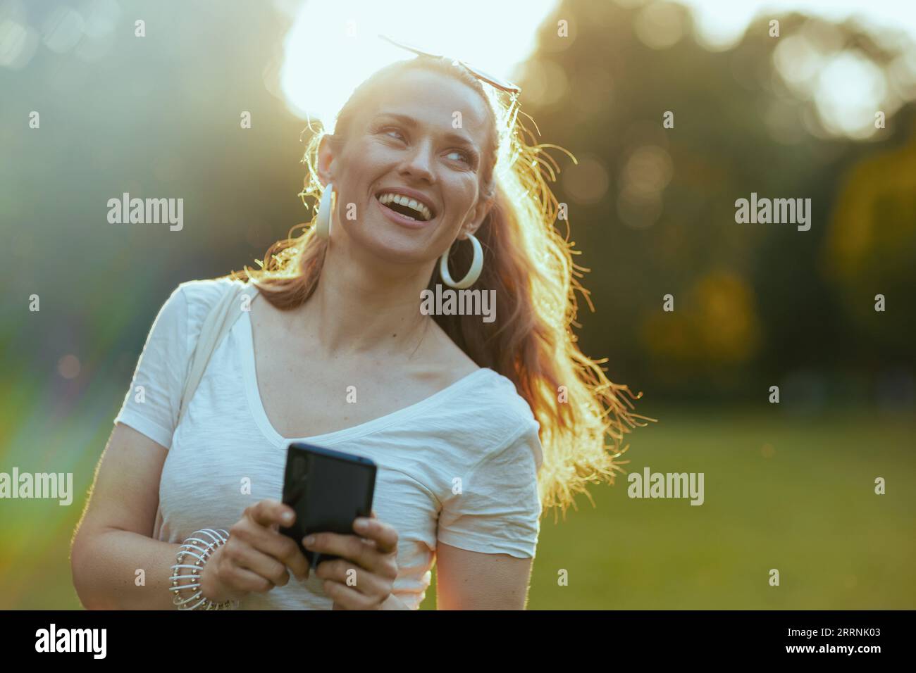 Heure d'été. heureuse femme d'âge moyen en chemise blanche en utilisant des applications de smartphone dans la prairie dans le parc de la ville. Banque D'Images