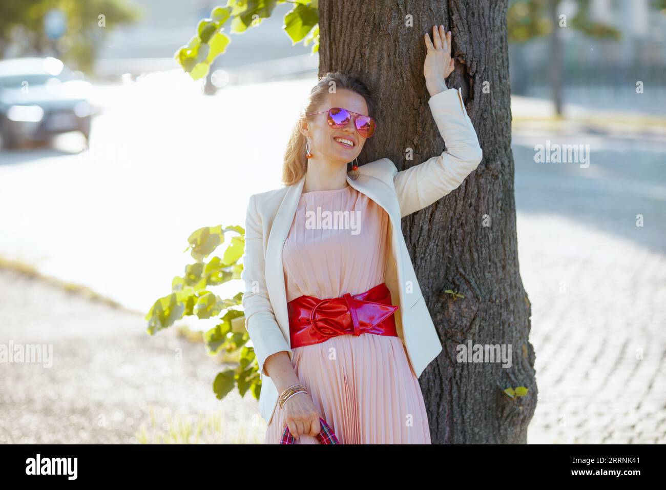 heureuse femme élégante de 40 ans en robe rose et veste blanche dans la ville avec des lunettes de soleil. Banque D'Images