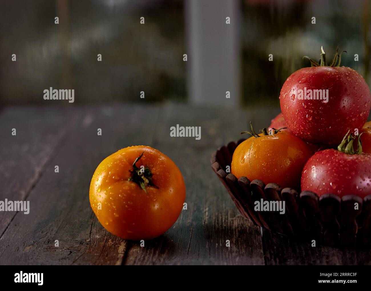 Tomates rouges et jaunes avec des gouttes de rosée dans un panier en bois sur une table en bois avec une fenêtre sombre en arrière-plan Banque D'Images