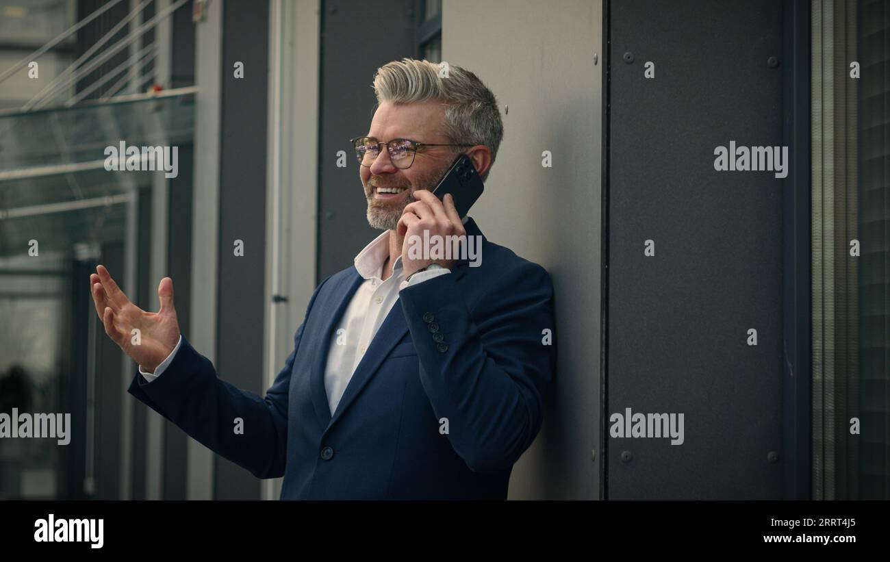 Entrepreneur d'employeur caucasien émotionnel à l'extérieur vieux homme d'affaires caucasien mature penchant à mur immeuble de bureaux souriant négocier smartphone Banque D'Images