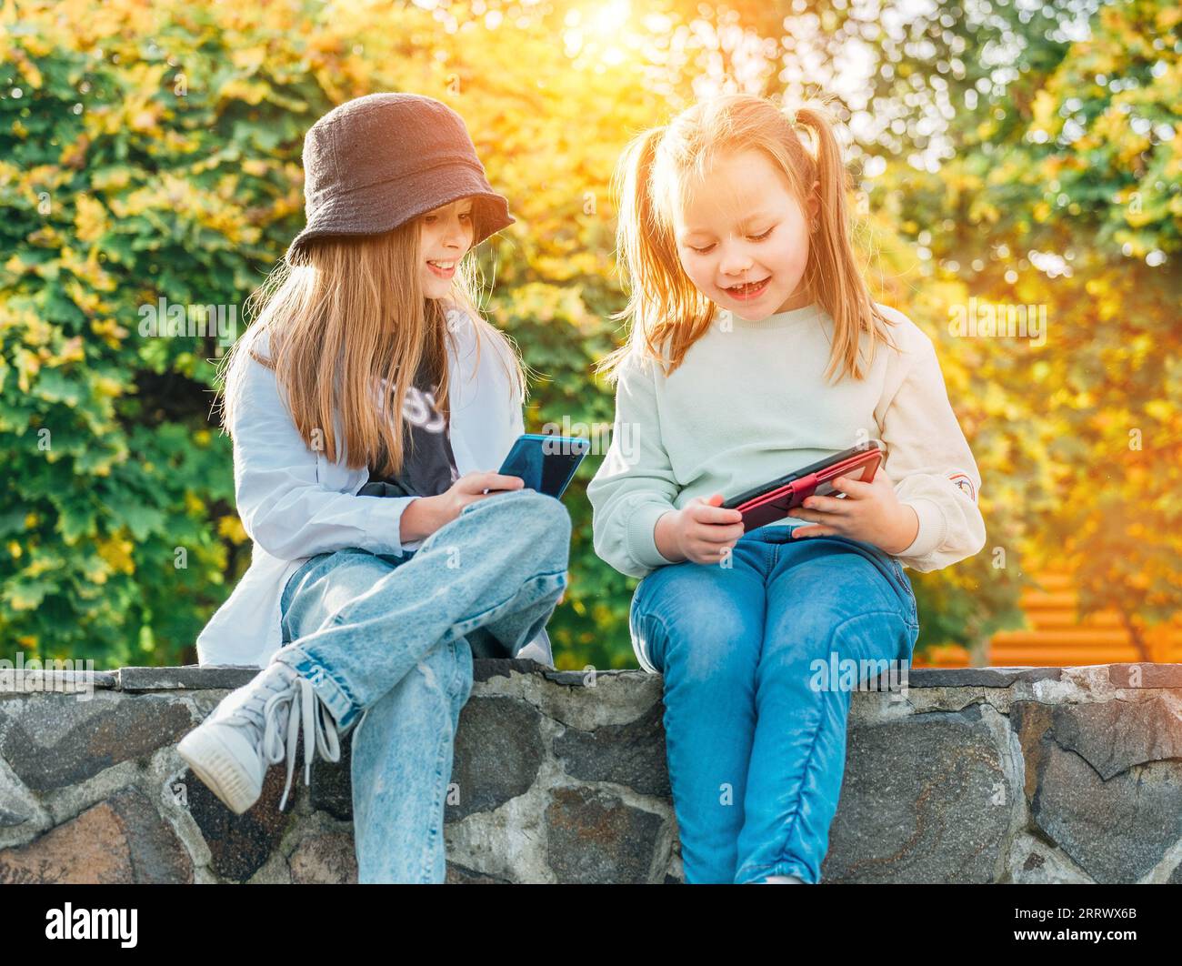 Deux petites filles sœurs souriantes, des enfants assis et parcourant leur smartphone dans le parc automnal. Une petite enfance imprudente et une te moderne Banque D'Images