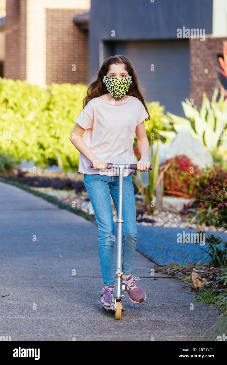 Une jeune fille caucasienne joue avec son scooter dans la rue par une soirée ensoleillée en Australie tout en portant un masque COVID Banque D'Images