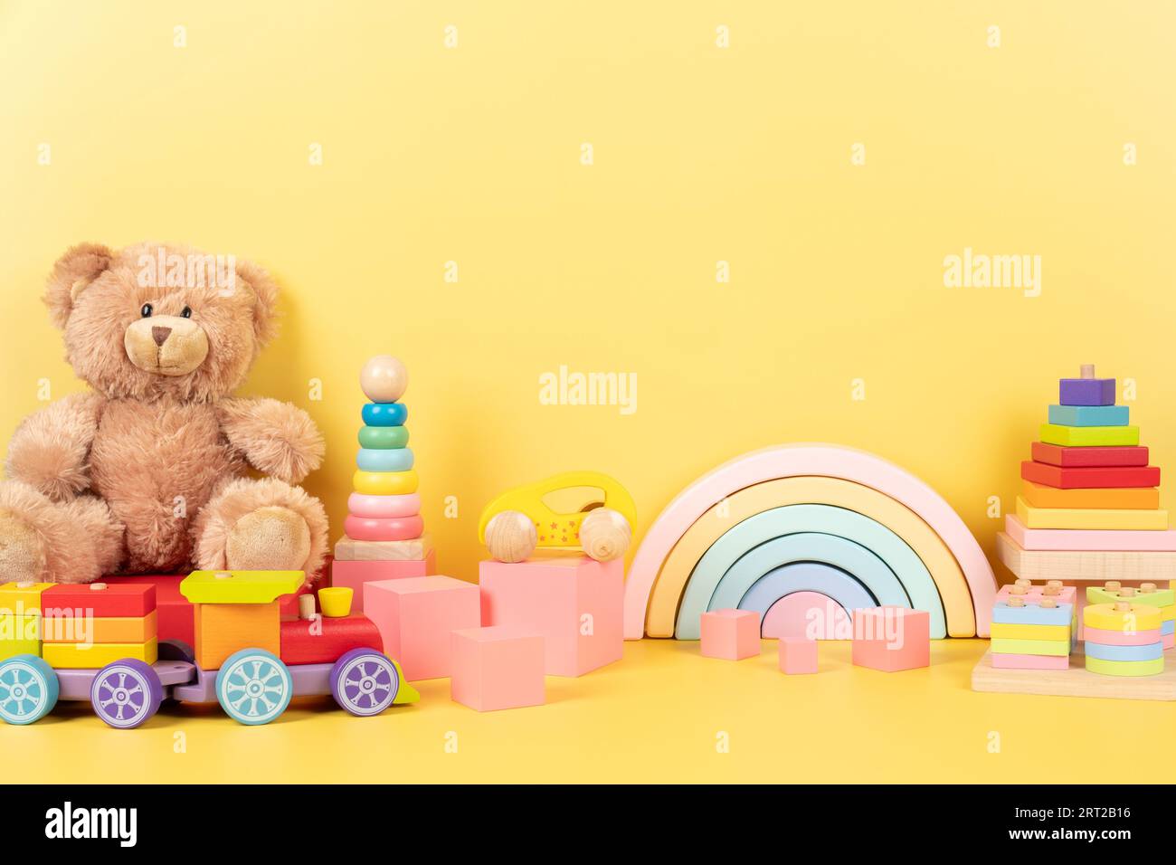 Collection de jouets éducatifs pour enfants. Ours en peluche, arc-en-ciel en bois, train, empilant des cubes roses pyramidaux sur fond jaune. Vue avant Banque D'Images